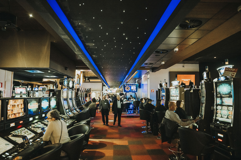 Une salle de casino animée avec des rangées de machines à sous de chaque côté de l'allée centrale. On voit plusieurs personnes qui sont assises sur des chaises noires en train de jouer aux machines.