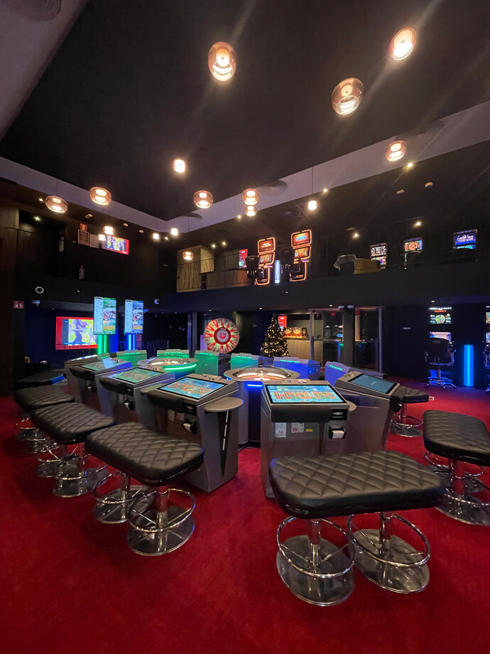 Une salle du casino d'Arlon avec un tapis rouge. On voit plusieurs tables de jeu électroniques sont qui sont disposées au centre, chacune équipée de sièges noirs rembourrés.