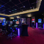 Une salle du casino d'Aubange avec un tapis rouge orné de motifs noirs. Des machines à sous et des bornes de jeux électroniques sont alignées le long des murs.