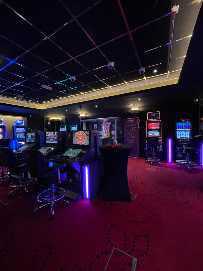 Une salle du casino d'Aubange avec un tapis rouge orné de motifs noirs. Des machines à sous et des bornes de jeux électroniques sont alignées le long des murs.