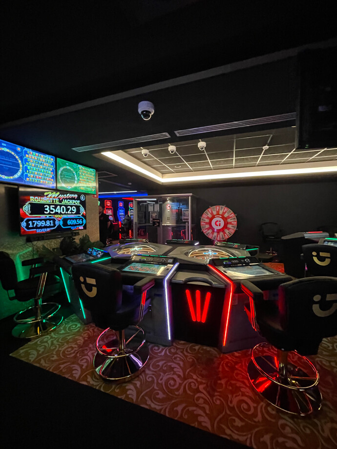 Salle de casino de Couvin avec un éclairage tamisé et des lumineux rouges et verts. Au centre, une table de jeu électronique équipée de sièges noirs.