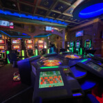 Salle du casino de Gosselies avec un plafond décoratif et un éclairage bleu. Des machines à sous sont alignées le long des murs, et des tables de roulette électroniques sont au centre de la pièce.