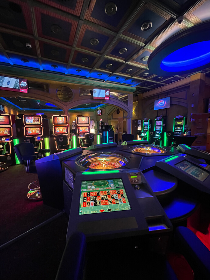 Salle du casino de Gosselies avec un plafond décoratif et un éclairage bleu. Des machines à sous sont alignées le long des murs, et des tables de roulette électroniques sont au centre de la pièce.