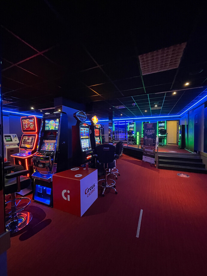 Une allée du casino de Grobbendonk avec des machines à sous alignées de chaque côté et l'éclairage est bleu et rouge.