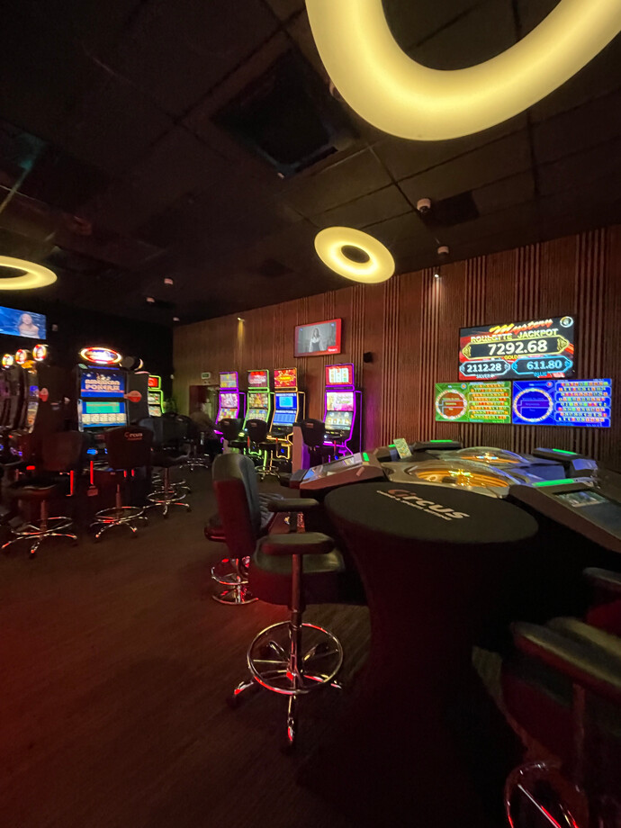 Salle de casino de Havay avec des anneaux lumineux suspendus au plafond. Des rangées de machines à sous colorées et des tables de jeu électroniques sont réparties dans l'espace.