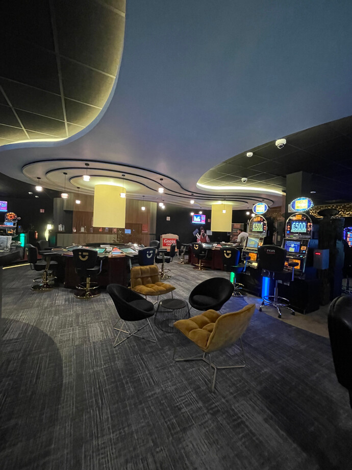 Salle de casino de Herstal avec un plafond incurvé et un éclairage moderne. Des sièges noirs et dorés sont disposés autour des tables de jeu.