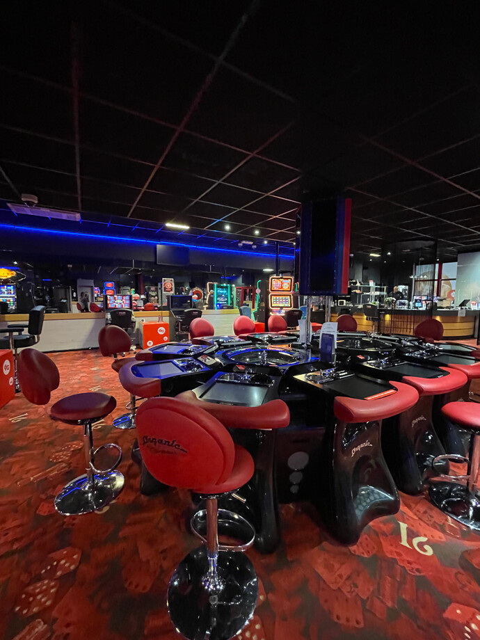 Salle de casino de Lier avec un sol rouge orné de motifs de cartes. Au centre, une table de jeu circulaire avec des écrans et des sièges rouges.