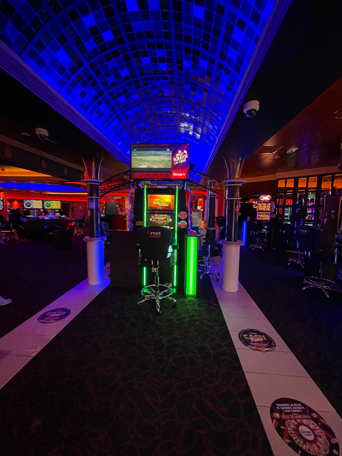 Une salle du casino de Loverval avec un plafond voûté éclairé en bleu. Des machines à sous et des bornes de jeu sont disposées de chaque côté de l'allée centrale. L'éclairage est bleu et rouge.