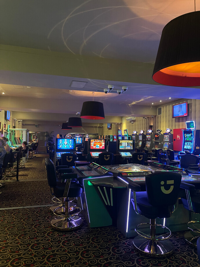 La grande salle de casino de Quievrain avec des machines à sous modernes. Les sièges devant les machines sont noirs avec des accents lumineux bleus. Le sol est recouvert d'un tapis à motifs circulaires.