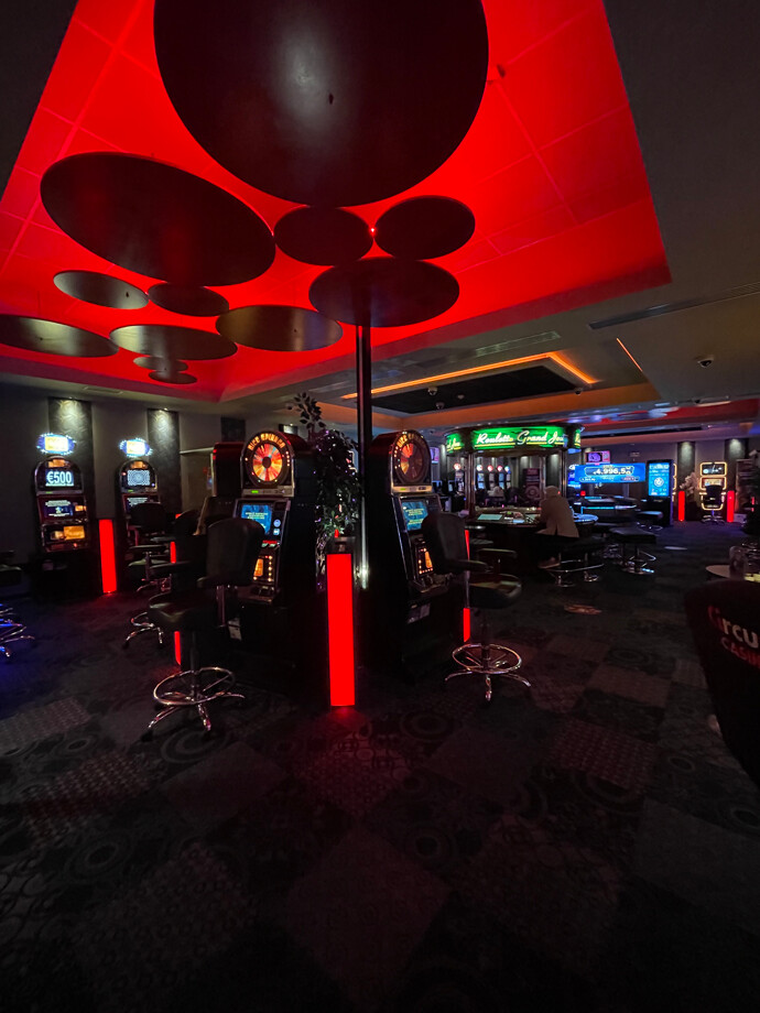 Vue de la salle de casino de Rocourt avec un plafond décoratif éclairé en rouge. Au centre, il y a des machines à sous entourées de chaises noires.