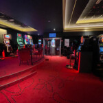 La salle de casino de Verviers avec un tapis rouge orné de motifs noirs. Il y a plusieurs machines à sous alignées le long des murs, éclairées par des lumières LED colorées.