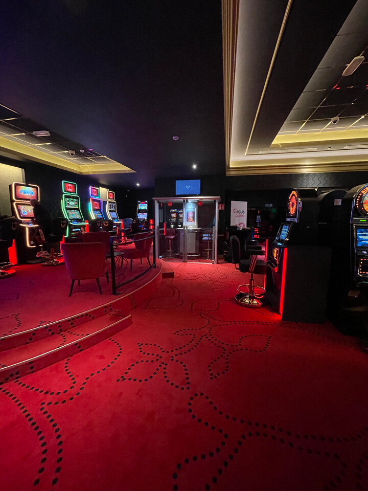 La salle de casino de Verviers avec un tapis rouge orné de motifs noirs. Il y a plusieurs machines à sous alignées le long des murs, éclairées par des lumières LED colorées.